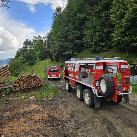 Ein roter Feuerwehrwagen mit der Aufschrift 'Feuerwehr Mixnitz' ist auf einem Feldweg geparkt. Dahinter steht ein kleinerer roter Wagen, und links steht ein Stapel Holz.