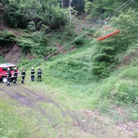 Feuerwehrleute in voller Ausrüstung bereiten sich darauf vor, ein rotes Boot von einem Felsvorsprung mit Seilen zu heben. Das Boot ist über einem steilen Hang mit dichter Vegetation suspendiert.