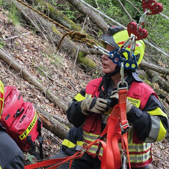 Ein Retter in einem gelben Helm und roter Weste mit Seilen und Gurten, bereit zur Unterstützung einer Person im Wald.