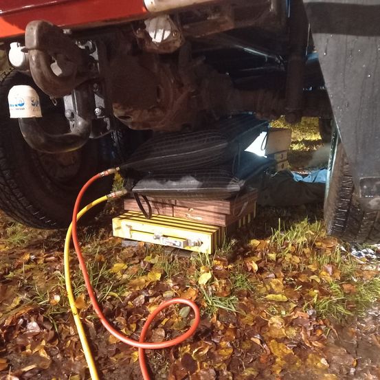 Eine Nahaufnahme des Unterbodens eines Fahrzeugs zeigt ein gelbes Gerät mit angeschlossenen Kabeln, das auf dem Boden zwischen gefallenen Blättern liegt.