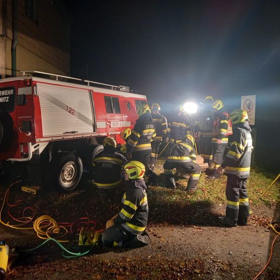 Eine Gruppe von Feuerwehrleuten ist um einen roten Feuerwehrwagen in der Nacht versammelt, einer von ihnen kniet.