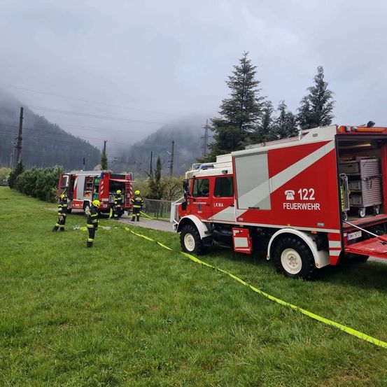 Drei Feuerwehrleute in gelben Helmen stehen neben einem Feuerwehrauto mit der Nummer 122 auf einer Rasenfläche, im Hintergrund befindet sich eine Bergkette.