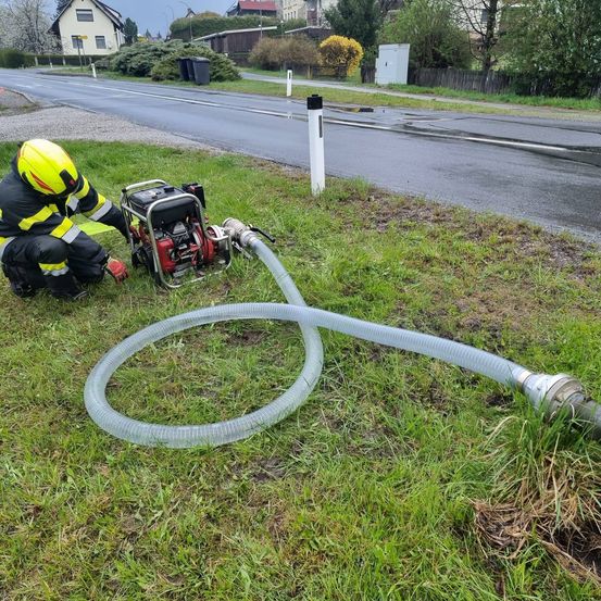 Ein Feuerwehrmann kniet am Straßenrand und arbeitet mit einer Pumpe und einem Schlauch in der Nähe eines Grasbereichs. Eine nasse Straße und Wohngebäude sind im Hintergrund zu sehen.