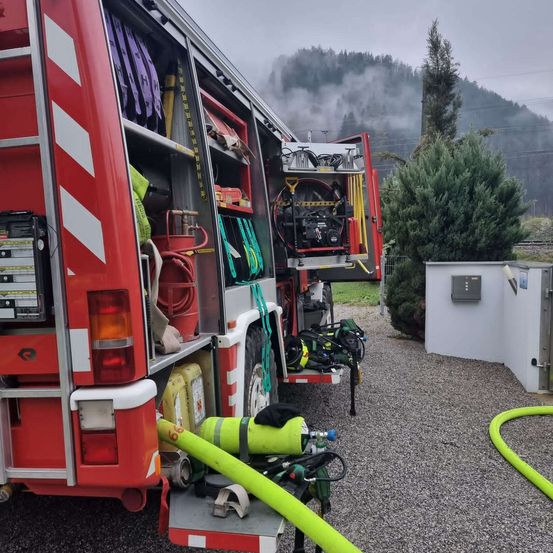 Ein roter Feuerwehrwagen mit offenen Abteilen, einem gelben Schlauch und verschiedenen Ausrüstungsgegenständen steht auf einem Schotterplatz. Dahinter sind ein grüner Baum und eine weiße Struktur zu sehen.