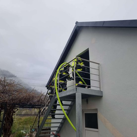 Feuerwehrleute in gelben Uniformen klettern eine Treppe hinauf, die zu einem Hauseingang führt. Sie halten einen gelben Schlauch. Das Haus hat ein Metallgeländer, eine weiße Tür und eine graue Wand.