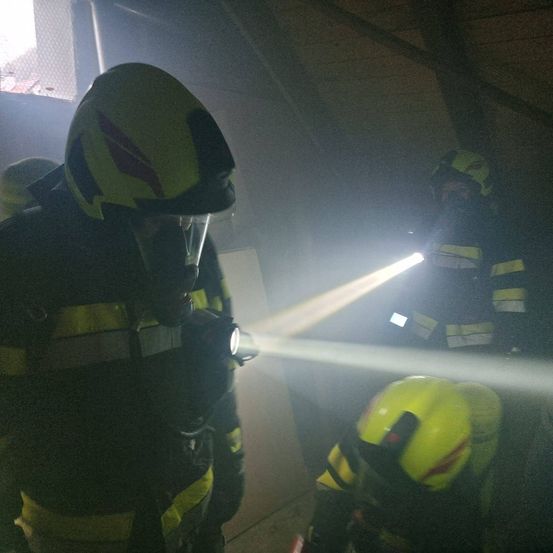 Feuerwehrleute in Schutzkleidung sind in einem rauchigen Raum. Einer benutzt eine Taschenlampe, während ein anderer hinter ihm steht. Der Raum ist schwach beleuchtet.