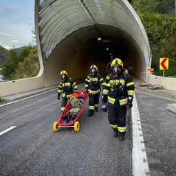 Drei Feuerwehrleute in Schutzkleidung schieben eine Trage durch einen Tunnel. Die Trage wird von einem roten Wagen mit gelben Rädern gezogen.