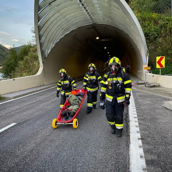 Drei Feuerwehrleute in Schutzkleidung schieben eine Trage durch einen Tunnel. Die Trage wird von einem roten Wagen mit gelben Rädern gezogen.