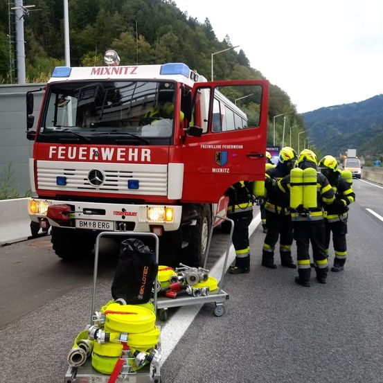 Ein Feuerwehrfahrzeug ist am Straßenrand geparkt, um das Feuerwehrleute herumstehen. Der Wagen ist mit 'Feuerwehr' beschriftet und hat ein Nummernschild 'BM 8 ERG'. Ausrüstung und Taschen befinden sich auf einem Trolley neben dem Fahrzeug.