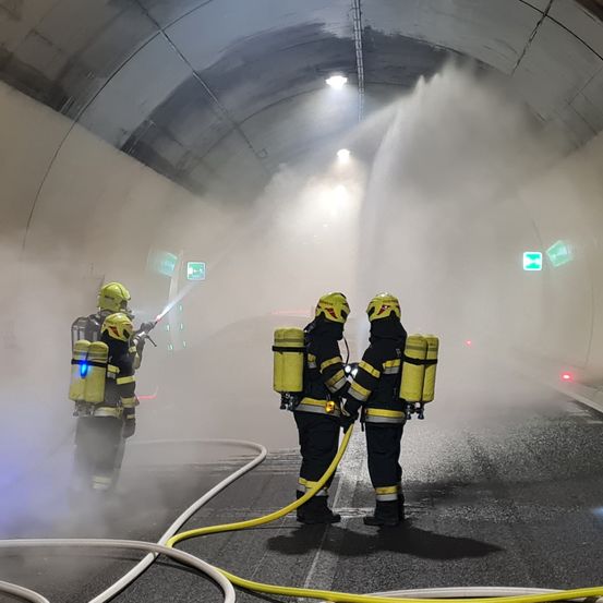 Drei Feuerwehrleute stehen in einem Tunnel und sprühen mit Schläuchen Wasser. Sie tragen Helme und Schutzkleidung. Der Tunnel ist mit Rauch gefüllt.
