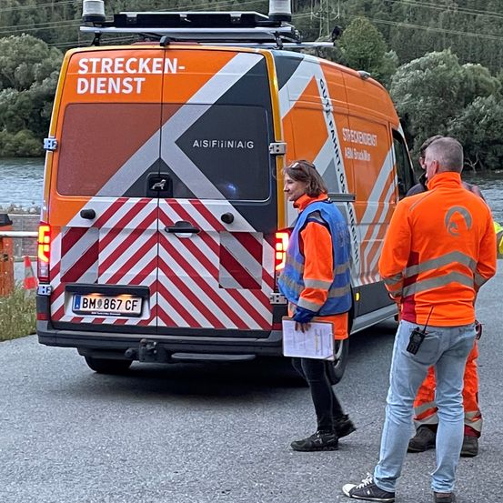 Zwei Arbeiter in orangefarbenen Uniformen stehen vor einem Van mit der Aufschrift 'Strecken-Dienst' und dem Kennzeichen 'BM 867 CF'. Eine Frau hält eine Clipboard, während ein Mann neben ihr steht. Dahinter sind ein Fluss und Bäume zu sehen.