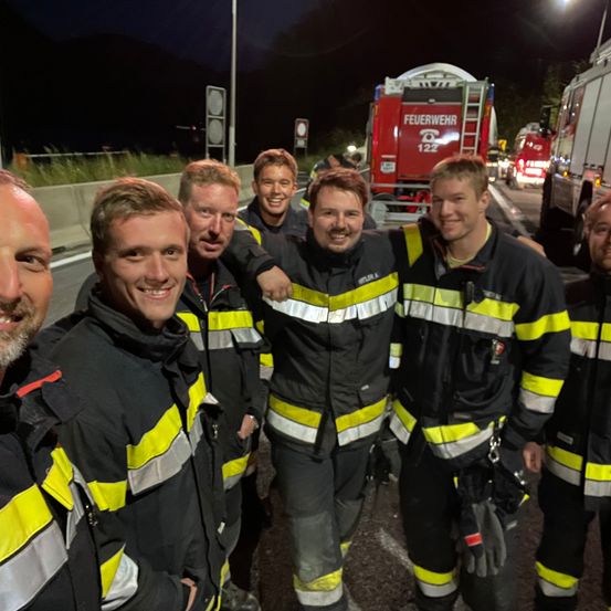 Eine Gruppe von Feuerwehrleuten in Uniformen steht nachts auf einer Straße und posiert für ein Foto mit einem Feuerwehrauto im Hintergrund.