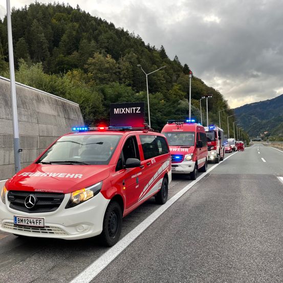 Rote Feuerwehrfahrzeuge mit blinkenden Lichtern sind am Straßenrand geparkt, mit Bergen und Bäumen im Hintergrund.