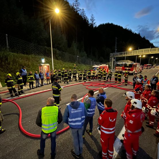 Eine Gruppe von Feuerwehrleuten in Uniformen steht nachts auf einer Straße, mit roten Schläuchen und Fahrzeugen in der Nähe. Ein Mann in einer gelben Weste spricht.