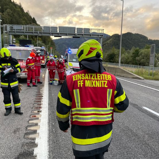 Rettungskräfte in Schutzkleidung stehen am Straßenrand, mit Bergen und einer Brücke im Hintergrund.
