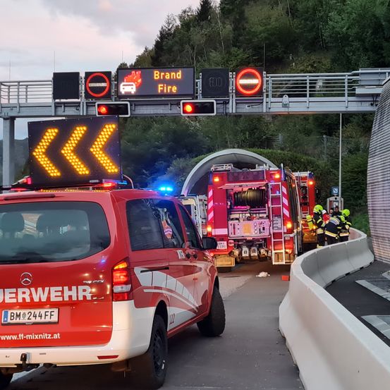 Einsatzfahrzeuge stehen am Tunneleingang. Ein Auto mit Polizeilichten ist in der Nähe. Über dem Tunnel warnen Verkehrsschilder vor einem Brand.