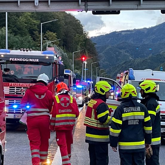 Einsatzkräfte in roten und schwarzen Uniformen stehen am Straßenrand, dahinter ein roter Feuerwehrwagen und ein weißer Krankenwagen. Ein Verkehrslicht befindet sich am Straßenrand, und Berge sind in der Ferne.