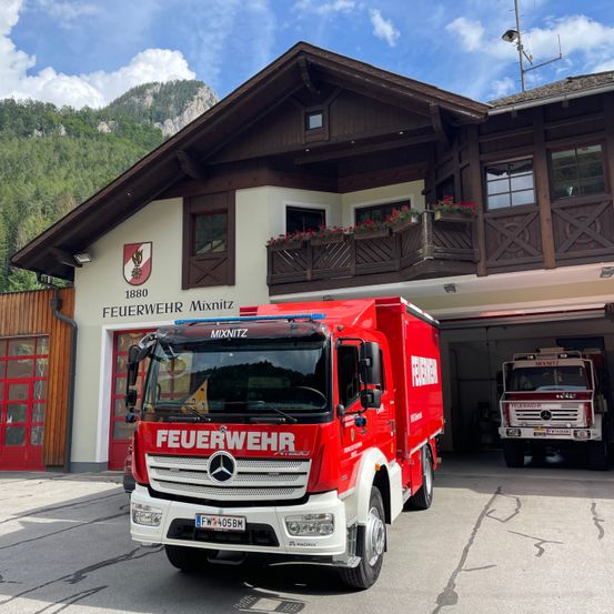 Ein roter Feuerwehrwagen steht vor dem Feuerwehrhaus Mixnitz, ein weiterer Feuerwehrwagen befindet sich in der Garage, vor einem Bergpanorama.
