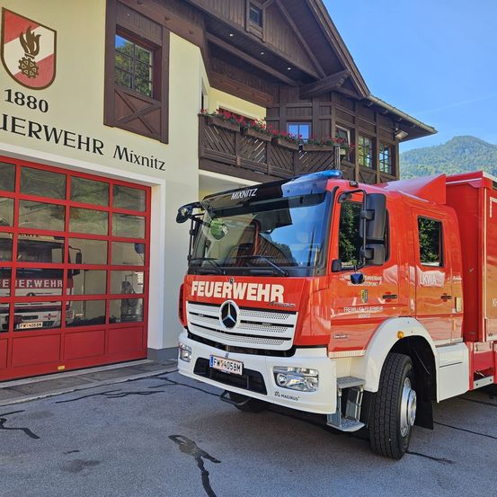 Ein roter Feuerwehrwagen steht vor einem Feuerwehrhaus mit dem Namen Mixnitz und einem Logo. Der Wagen hat ein Nummernschild und ist am Straßenrand geparkt.
