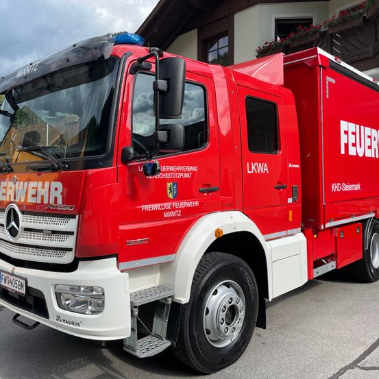 Ein roter Feuerwehrwagen mit dem Wort 'FEUER' auf der Seite steht vor einem Gebäude. Der Wagen ist von der Marke LKW und hat ein Nummernschild.