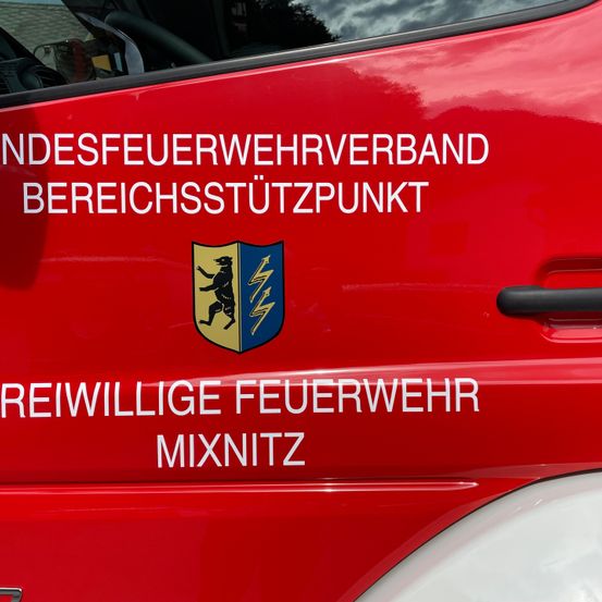 Ein rotes Fahrzeug mit dem Text 'NDSFEUERWERWERBAND BEREICHSSSTUTZPUNKT' und 'REIWILLIGE FEUERWEHR MIXNITZ' unter einem Schild mit einem Bären und Blitz.