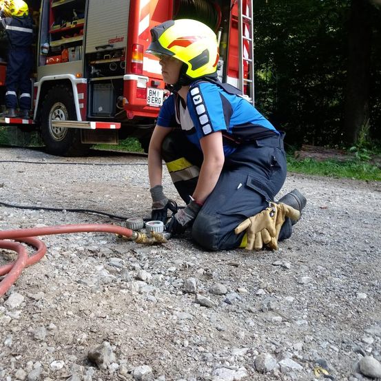 Ein Feuerwehrmann kniet auf dem Boden und hält eine Schlauchkupplung. Dahinter steht ein roter Feuerwehrwagen, der offen ist, und ein weiterer Feuerwehrmann steht darin.