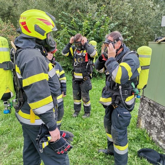 Feuerwehrleute in gelben und grauen Uniformen stehen auf einem Rasen, sie stellen ihre Helme und Masken ein. Bäume sind im Hintergrund zu sehen.