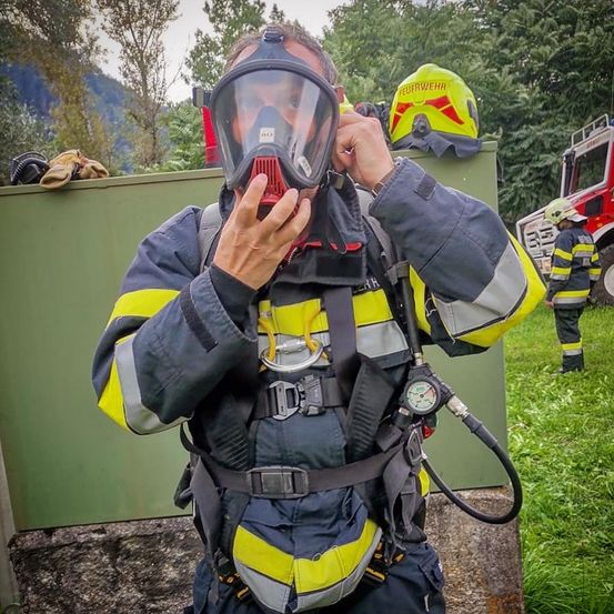 Ein Feuerwehrmann justiert seine Maske, während er ein gelb-graues Uniform mit reflektierenden Streifen trägt. Ein Feuerwehrhelm ruht auf einer grünen Struktur hinter ihm.
