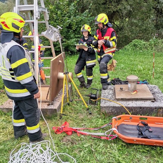 Eine Gruppe von Feuerwehrleuten in Schutzausrüstung steht im Freien, möglicherweise in Ausbildung. Einer hält ein Tablet, während ein anderer eine Struktur beobachtet. In der Nähe befinden sich eine Leiter, Seile und verschiedene Ausrüstungsgegenstände.