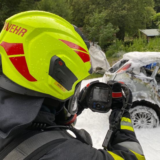 Ein Feuerwehrmann fotografiert ein beschädigtes Fahrzeug mit einer Kamera. Der Feuerwehrmann trägt einen gelben Helm mit roten Streifen.