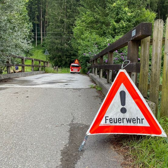 Ein roter Feuerwehrwagen fährt auf einer Straße mit einem Warndreieck auf der Seite. Im Hintergrund sind Bäume und ein Holzzaun zu sehen.