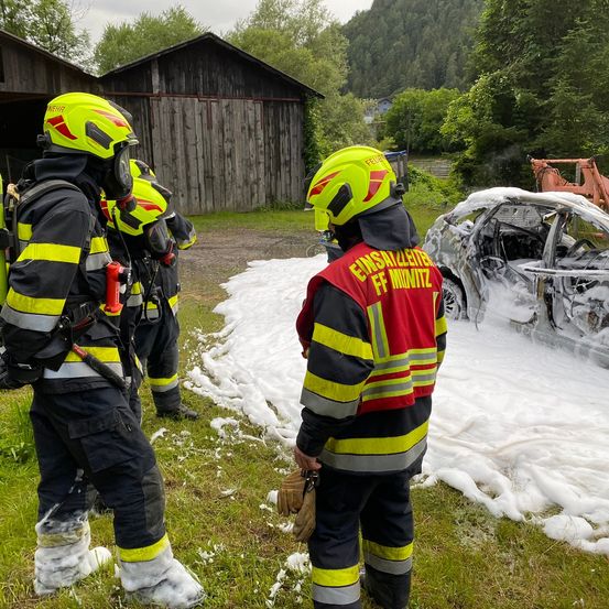 Feuerwehrleute in gelben Helmen stehen neben einem verbrannten Auto, das mit Schaum bedeckt ist. Dahinter steht eine Holzscheune und ein grünes Feld.