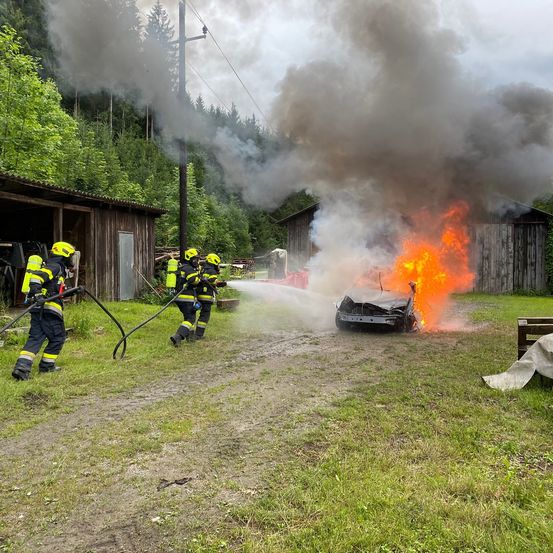 Feuerwehrleute löschen ein brennendes Auto in einem ländlichen Gebiet, wobei Rauch aus einem nahegelegenen Scheunen aufsteigt.