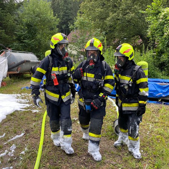 Drei Feuerwehrleute in gelben und schwarzen Uniformen mit Masken und Handschuhen stehen in einem Grasbereich mit einem gelben Schlauch in der Nähe. Bäume und ein Haus sind im Hintergrund.