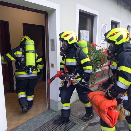 Drei Feuerwehrleute in Schutzkleidung verlassen ein Gebäude, einer hält einen Feuerlöscher. Sie stehen auf einem gefliesten Boden.