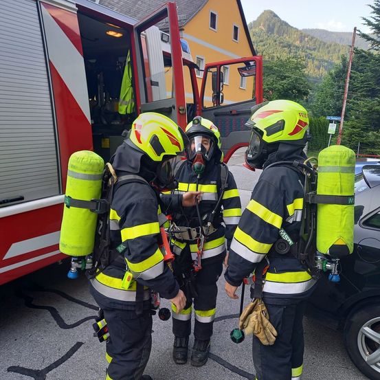 Drei Feuerwehrleute mit gelben Helmen und Sauerstoffflaschen stehen vor einem Feuerwehrwagen mit offener Tür. Die Szene spielt sich draußen ab, mit einem gelben Gebäude und Bergen im Hintergrund.