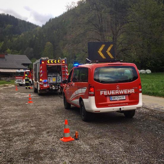 Ein Feuerwehrfahrzeug ist auf einem Schotterplatz geparkt, mit einer Person in der Nähe. Ein orangefarbener Verkehrskegel ist in der Nähe. Ein weiteres Fahrzeug steht dahinter.