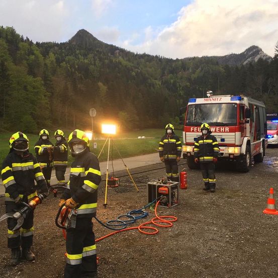 Feuerwehrleute in Schutzkleidung bereiten Ausrüstung in der Nähe eines Feuerwehrwagens vor, mit Bergen im Hintergrund.