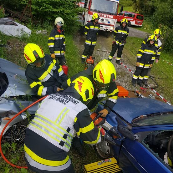 Feuerwehrleute arbeiten an einem Autounfall mit zwei beschädigten Fahrzeugen. Sie verwenden Werkzeuge und Ausrüstung an den beschädigten Autos auf einem Grasfeld.