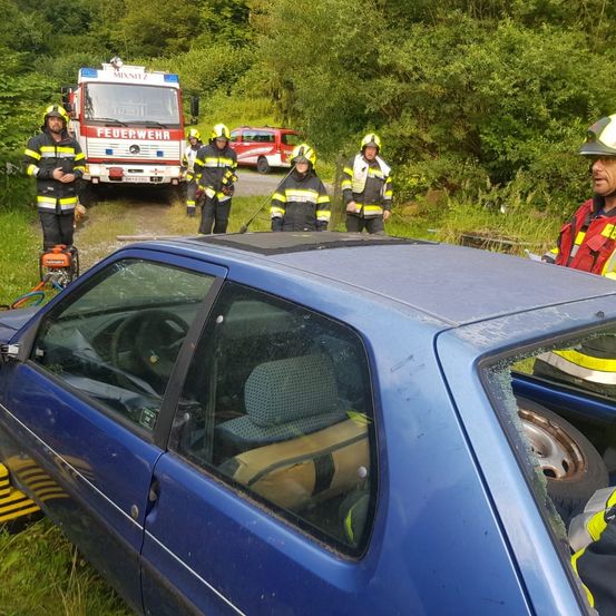 Feuerwehrleute in Uniform stehen neben einem demoliert blauen Auto. Ein Feuerwehrwagen ist hinter ihnen geparkt, und ein rotes Auto ist im Hintergrund sichtbar.