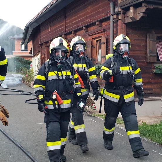 Drei Feuerwehrleute gehen mit Schutzausrüstung und Masken auf einem Gehweg. Sie werden von einem vierten Feuerwehrmann begleitet, der teilweise sichtbar vor einem Holzhaus steht.
