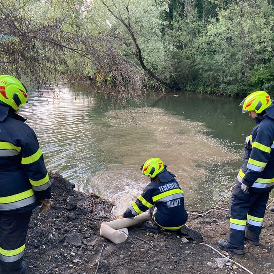 Drei Feuerwehrleute in voller Ausrüstung stehen am Flussufer, einer kniet und hält einen Schlauch in der Nähe des Wassers, während die anderen in der Nähe stehen.