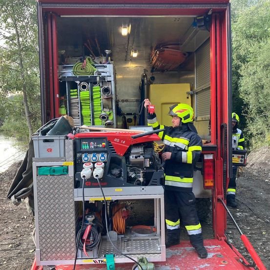 Ein Feuerwehrmann mit gelbem Helm steht in einem roten Lkw. Der Lkw ist mit verschiedenen Geräten und Werkzeugen gefüllt. Ein weiterer Feuerwehrmann steht in der Nähe.