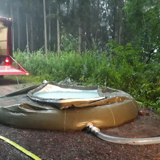 Ein aufblasbarer Pool liegt entfaltet am Boden, angeschlossen an einen Schlauch. Ein rotes Fahrzeug ist in der Nähe. Die Szene spielt sich in einem Wald mit dichter Vegetation ab.