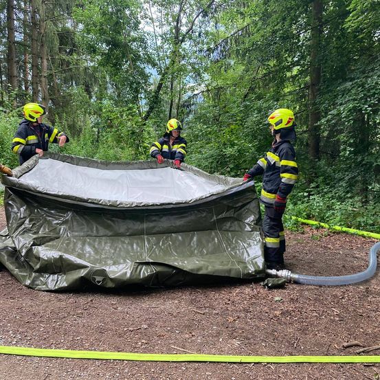 Drei Feuerwehrleute in gelben Helmen bauen ein grünes Planenzelt in einem Wald auf. Sie halten die Plane und einer sieht sich es an.