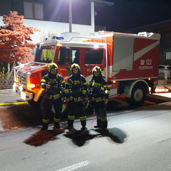 Drei Feuerwehrleute stehen vor einem Feuerwehrauto in der Nacht. Sie tragen vollständige Schutzausrüstung, einschließlich Helme und Stiefel. Das Feuerwehrauto ist am Straßenrand geparkt.