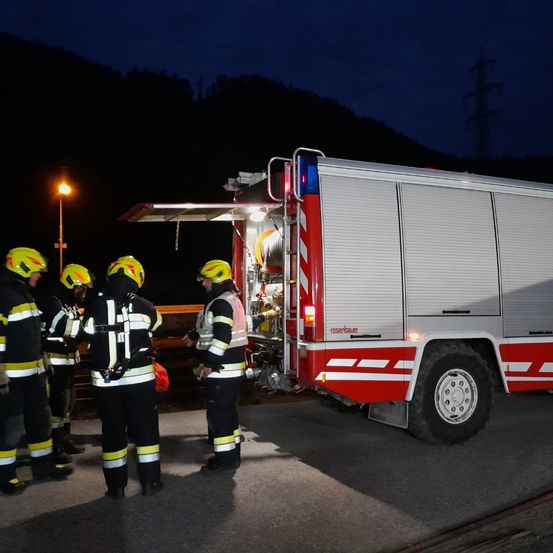Feuerwehrleute in Schutzkleidung stehen nachts um einen Feuerwehrwagen herum, wobei eine Lampe die Szene beleuchtet.