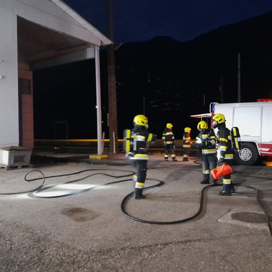 Feuerwehrleute in Schutzkleidung stehen bei einem Feuerwehrwagen und Schläuchen in der dunklen Nacht. Berge sind im Hintergrund sichtbar.
