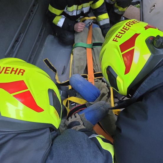Feuerwehrleute sichern eine Person auf einer Trage in einem Fahrzeug, die gelbe Helme mit der Aufschrift 'FEUERWEHR' tragen. Die Person ist mit orangefarbenen und grünen Gurten befestigt.