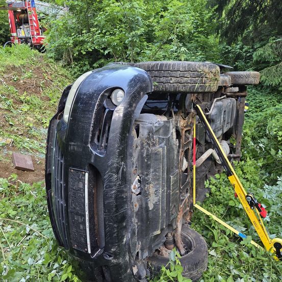 Ein umgekipptes Auto ruht in einem bewaldeten Gebiet auf der Seite, mit zwei noch befestigten Reifen. Der vordere Stoßfänger ist beschädigt und ein gelbes Gerät ist befestigt. Ein Feuerwehrwagen ist im Hintergrund zu sehen.